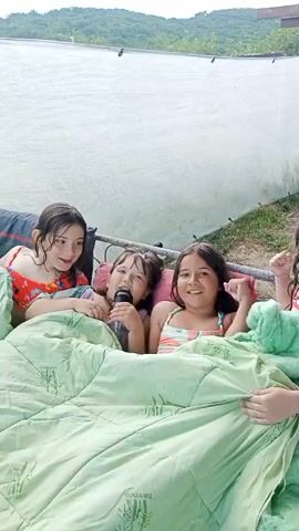 Four young girls are lying on their backs under a green blanket, which has a bamboo pattern printed on it. The girls are smiling and appear to be having fun. One girl in the middle is holding a black object, possibly a microphone, and is singing. Another girl on the right has her arm raised in the air. They are outside, under a white tarp that is stretched over a frame. In the background, there are rolling green hills and trees, suggesting a rural or natural setting. The sky is bright, indicating it is daytime, likely sunny. The overall atmosphere is one of joy and lightheartedness.