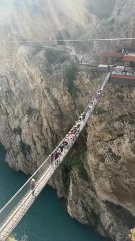 A high-angle shot shows a long suspension bridge spanning a deep canyon with steep, rocky walls. A bright turquoise river flows at the bottom of the canyon. A large group of people is walking across the bridge from the left side of the frame towards a multi-level structure with balconies visible on the far canyon edge. Most of the bridge is densely packed with pedestrians, while a single individual walks ahead of the main group near the camera's vantage point.