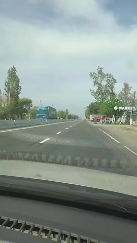 The view from the dashboard of a car traveling on a paved road with two lanes in each direction. There is a median on the left side and an unpaved shoulder on the right side. A woman in a long green dress is standing on the shoulder of the road waving her arms. Several trucks and cars are parked on the shoulder of the road. There are trees on both sides of the road, and streetlights are visible on the left side. The sky is mostly cloudy.