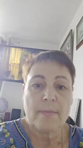 A video shows a woman recording herself in an indoor setting, likely a home in Cwmbrân, United Kingdom. The woman, who appears to be in her 50s or 60s, has short, light brown hair and a neutral expression as she looks directly at the camera. She is wearing a blue top with a patterned neckline.

In the background, a flat-screen television is mounted on a white wall. The TV displays a person with dark skin and dark hair, who appears to be engaged in a phone call or holding an object to their ear. To the right of the TV, several framed pictures are hanging on the wall.

Below the TV, a surface, possibly a table or shelf, holds various items, including a small globe and what looks like a tablet or book. Part of a colorful, possibly knitted or crocheted, blanket or throw is visible in the bottom left corner of the frame. The overall lighting suggests it is daytime. No discernible text is visible in the background. The video includes audio of spoken words in an unfamiliar language.