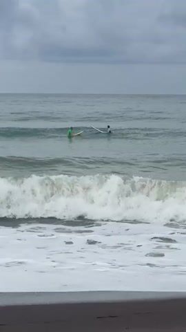 The video shows a segment of a beach with a gray, cloudy sky overhead. The ocean is choppy with moderate waves breaking near the shore, leaving a foamy white line on the dark sand. Two people, each with a surfboard, are visible in the water further out. One person is wearing a green top, and the other is wearing a dark top. They appear to be waiting for waves or possibly socializing. The water is a muted gray-green color. The camera is positioned on the beach, looking out towards the ocean. The waves are the most dynamic element in the foreground.