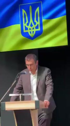 A man in a light brown suit with a white shirt and glasses stands at a wooden podium with a microphone. He appears to be speaking. A large screen behind him displays the Ukrainian flag. The backdrop behind the speaker is black.