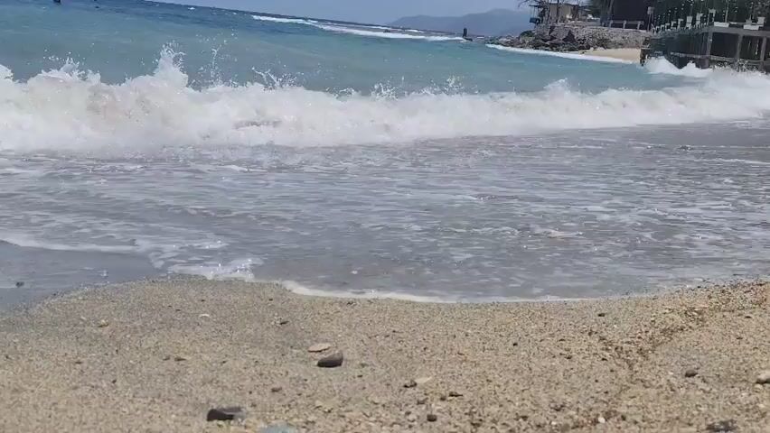 This video captures a close-up view of a sandy beach with waves gently washing ashore. The water is a clear turquoise, transitioning to white foam as the waves break. Small pebbles and shells are scattered on the wet sand. In the background, the ocean extends to the horizon under a bright sky, and hints of land with buildings and vegetation are visible. The camera appears to be positioned very low to the ground, giving a unique perspective of the water's edge.