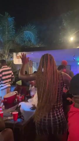 The video shows an outdoor gathering at night. Several people are visible, some dancing and others sitting at tables. There are red cups and bottles on the tables, suggesting a party or social event. String lights and blue lighting illuminate the scene. A woman with long braided hair is in the foreground, facing away from the camera with her arm raised. In the background, a person in a striped shirt is also visible. The environment appears to be a patio or garden area with trees and greenery.
