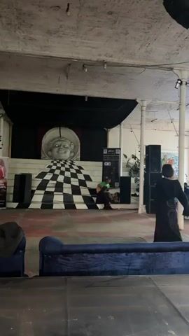 The video opens on a large, dimly lit indoor space with a stage in the background. A large, white sculpture of a face with wide eyes and a smile is positioned at the top of a set of stairs. The stairs are painted with a black and white checkered pattern, creating a disorienting effect. A person in a black outfit is standing and facing away from the camera, looking towards the stage. Another person is sitting on the stairs playing a guitar. There are speakers on either side of the stage. The environment appears to be a performance or art space with a somewhat industrial aesthetic, evidenced by the exposed pipes and concrete ceiling.