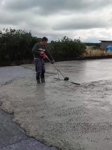 A man is shown operating a screed, a tool used to level wet concrete. He is standing in a large, outdoor expanse of wet concrete, wearing work clothes and rubber boots. The concrete appears to be freshly poured, with a rough texture on the edges and a smoother, wet surface in the center where the man is working. The background shows greenery, a fence, and a cloudy sky. The man is holding the screed with both hands, guiding it across the concrete surface. The screed has a long handle and a flat blade that smooths and levels the concrete.