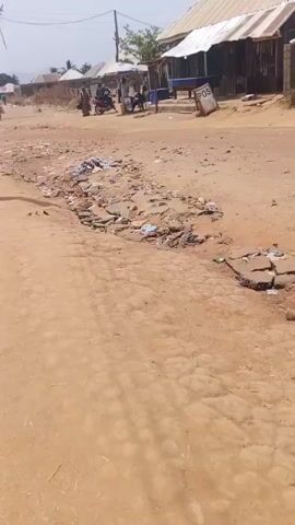 The video shows a dirt road with broken pavement and litter. Several people on motorcycles pass by in the background. In the background are buildings and a sign that says "POS". The road is in disrepair with chunks of pavement broken and scattered. There is trash and debris visible among the broken pavement.