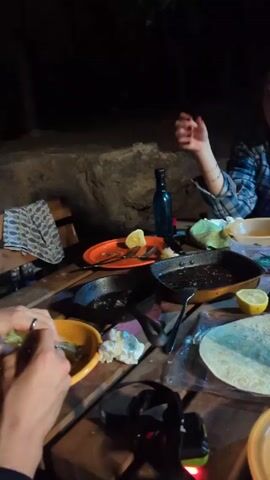 Here is a description of the video:

Two individuals are seen at a wooden table outdoors at night, preparing food. On the table are various cooking utensils, including two frying pans, plates, a bottle of blue liquid, tortillas, and a lemon. One person is assembling a taco, while the other reaches for something in one of the pans. The setting is dimly lit, with a large rock visible in the background.
