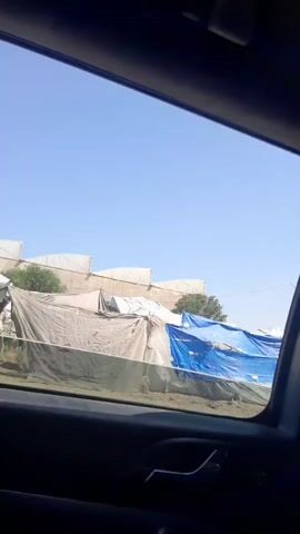 The video shows a view from inside a vehicle, likely a car, looking out the side window. The vehicle is moving, and outside the window, there is a makeshift settlement or camp. The structures appear to be constructed from various materials, including tarps of different colors (blue and white/grey) and possibly other salvaged items. The structures seem to be dwellings or shelters. The sky is clear and blue. There's a pole or thin tree trunk sticking up in the background. The scene conveys a sense of poverty and makeshift living conditions.