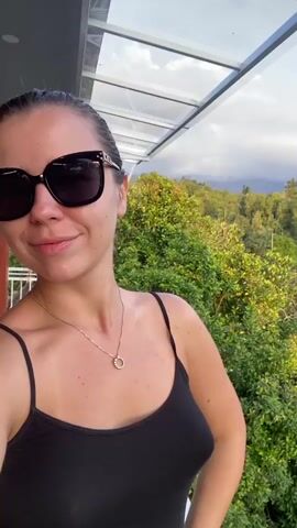 A woman with dark hair pulled back and wearing sunglasses is taking a selfie. She is wearing a black tank top and a silver necklace. She is standing on a balcony with a view of lush green trees and mountains in the background. The sky is cloudy.