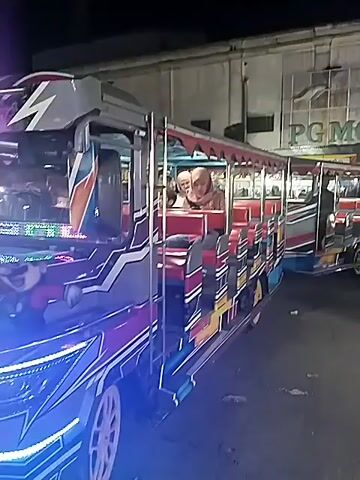 At night, the video shows a small train-like vehicle, brightly lit with blue and purple lights, at what appears to be an outdoor market or fair. Two people are seen riding in the vehicle. Several food stalls are visible, illuminated by bright lights. People, including adults and children, are walking around the area. One stall is labeled "JENANG". The overall atmosphere seems lively and festive.