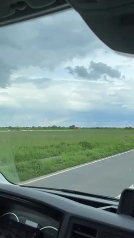 The video shows a rainbow in the distance, over a green field with a red building and power lines. The view is from inside a car driving on an asphalt road with a white line. The sky is cloudy. Two people are speaking in a language other than English.