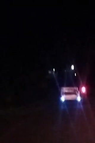 The video shows a car driving at night. The car's headlights are on, illuminating the dark road ahead. The taillights of the car are also visible, appearing red. There are some lights in the distance, possibly streetlights or other vehicles. The scene is dark and appears to be a rural or suburban area. There is no visible text in the video. The time of day is clearly night. The weather is not discernible. The location is identified as Keude Lueng Putu, Indonesia. There are no people clearly visible, and the focus is on the car and the driving experience at night. The overall mood is one of travel and perhaps solitude due to the darkness and lack of other apparent activity.