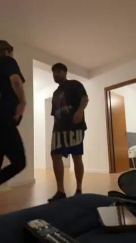In a medium shot inside what appears to be a home living room, a woman and a man are dancing together. The woman is on the left, wearing a black t-shirt, black leggings, and her long hair is partially tied back. The man is on the right, wearing a black t-shirt and blue shorts. They are holding hands and swaying to the music. In the foreground, a phone, a remote control, and other objects are visible. A television is on the left side of the shot, and a door is on the right side, leading to another room with a bed. The lighting is moderately bright.