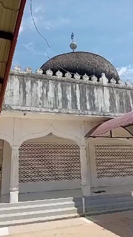 A low-angle, panning shot shows a mosque on a sunny day with a blue sky and scattered clouds. The mosque features a dark, dome-shaped roof with a crescent moon finial. The walls are weathered and discolored, with decorative crenellations along the top. A white, covered walkway with decorative arches and screens extends from the side of the mosque. Steps lead up to the walkway and the mosque entrance. The ground is paved with bricks in some areas and appears to be sandy in others. Two figures are visible in the background, one standing and another partially obscured.