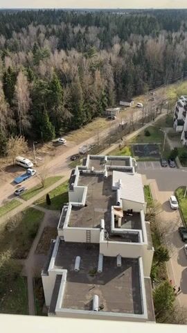 The video shows an elevated view of a residential area bordering a dense forest. Several cars are parked or driving on a dirt road that runs along the edge of the woods. A few small, utility-type vehicles are also visible, along with a few small buildings or sheds. To the right of the road is a multi-story apartment building with a flat, multi-tiered roof. There are patches of green grass and trees around the base of the building and a small playground or recreational area with a few pieces of equipment. A person, dressed in red, is visible walking near the building. The sky is overcast, and the lighting suggests it is daytime. The focus is on the scene and the interaction between the built environment and the natural landscape.