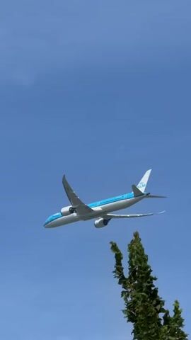 A KLM airplane flies across a bright blue sky. The airplane has a white body with blue and white wings and tail. The KLM logo is visible on the tail. The airplane is flying from left to right across the frame, with its nose slightly pointed upward. There are a few wispy white clouds visible in the lower part of the frame.