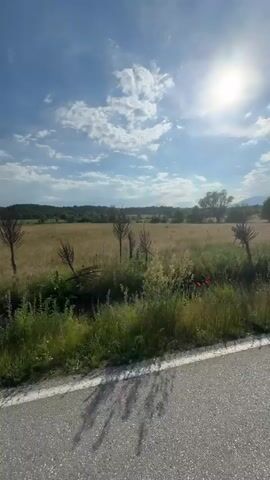 A first-person perspective video from a vehicle moving on a road in a rural area. The camera points slightly left and then pans to the center, showing the road, green grass and fields on both sides, and trees in the distance under a blue sky with some white clouds. The vehicle's dashboard is visible from the 00:03 mark.