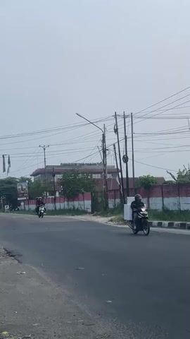 A vertical video shows a road with motorcycles and a building in the background. The road is paved and has a white line marking the edge. Several motorcycles are visible, some carrying passengers. In the background, there is a building with a red and white striped wall in front of it. Overhead, there are numerous power lines. The sky is overcast.