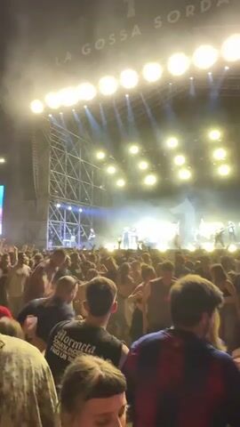 The video captures a crowd of people at a live music concert, illuminated by stage lights. A large scaffolding structure frames the stage, displaying the words "LA GOSSA SORDA".  People are closely packed together, moving and dancing to the music. Some individuals are clearly visible while others remain in shadow. The camera angle is from within the crowd, providing a sense of immersion in the concert atmosphere. The stage lights are bright and dynamic, adding to the energy of the scene. A pink inflatable figure can be seen on stage. A screen shows images of the band as they perform. Overall, the video conveys the excitement and vibrancy of a live music event.