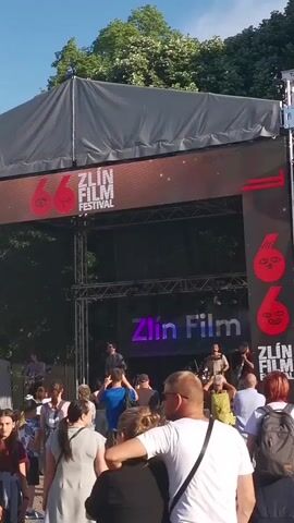 The video shows a crowd of people watching a band perform at an outdoor event. The stage is set up for the Zlín Film Festival, indicated by the banners and signage.  A large screen displays a close-up of the lead singer playing guitar. People are casually dressed and appear to be enjoying the music. Trees and a blue sky form the backdrop.