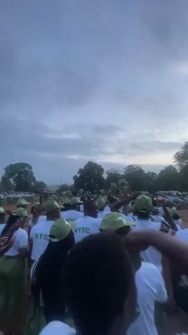 This media file captures a large gathering of people, predominantly young adults, in an outdoor setting. The subjects are dressed in matching uniforms consisting of white t-shirts with "NYSC" printed on the back and green caps. They appear to be participating in an event, possibly a ceremony or assembly, as they are arranged in rows and sections.

The scene is set in a somewhat rural or semi-urban area, indicated by the presence of trees, dusty ground, and what looks like a rudimentary structure or shelter in the background. The time of day is either early morning or late afternoon, suggested by the soft, diffused light and the overcast, cloudy sky. The weather appears to be mild, with no direct sunlight.

The activity seems to be a formal gathering or parade, with many individuals looking in the same direction, suggesting they are observing something or someone. Some people in the foreground are more clearly visible, and their actions suggest a mixture of participation and observation. One person is holding up a green object, possibly a leaf or a cap, while another is seen taking a picture with a mobile phone.

The overall mood is one of anticipation and engagement. The presence of the "NYSC" logo strongly suggests that this event is related to the National Youth Service Corps in Nigeria, a mandatory program for recent graduates. The location context of Emure-Ekiti, Nigeria, further solidifies this connection. The sky is a prominent feature, with dramatic cloud formations adding to the atmosphere of the scene.