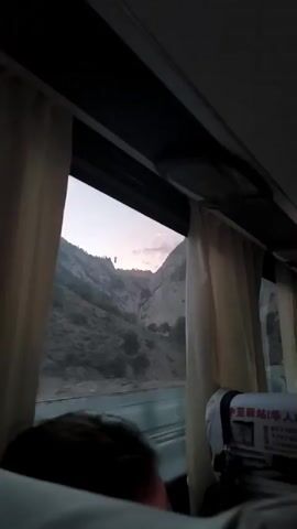 The video is a first-person perspective from inside a moving vehicle, likely a bus, looking out a window. The window is partially obscured by beige curtains on the left and right sides. Through the window, a mountainous landscape is visible, with steep, rocky slopes. The sky appears to be either dawn or dusk, with soft, pastel colors. In the foreground, a person's head and shoulder are visible, facing away from the camera and towards the window. Another person's head is visible further back in the vehicle. On the right side of the window, a seat pocket with some visible Chinese text is attached to the back of a seat. The vehicle is moving, indicated by the motion blur of the passing scenery. At one point, a person in the foreground holds up a smartphone, appearing to record the view. Another person's voice is heard saying "Don't film."