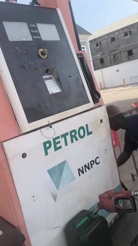 A short video shows a gas station attendant filling a green jerrycan with petrol from a gas pump. The pump is branded with “PETROL” and “NNPC”. The attendant is wearing a navy blue uniform with green accents. There are other people visible in the background, including another attendant and a person on a motorbike. The setting is an outdoor gas station with a building in the distance.