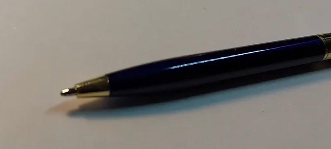 The video shows a close-up, static shot of a dark blue pen with gold accents lying on a light-colored surface. The pen is positioned horizontally, with the tip pointing to the left. The lighting creates reflections on the glossy surface of the pen, highlighting its metallic sheen and the gold trim around the tip and near the clip. The background is out of focus, suggesting a shallow depth of field. The video is brief and appears to be a still shot with no movement.