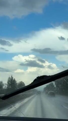 The video shows a view from inside a vehicle driving on a wet road. Raindrops are visible on the windshield. The road ahead is wet and reflects the sky. Other vehicles are also driving on the road. The sky is partly cloudy with patches of blue sky visible. Trees line the sides of the road. There are streetlights along the road. The lighting suggests it is daytime, possibly late afternoon. The overall atmosphere is one of driving in rainy weather.