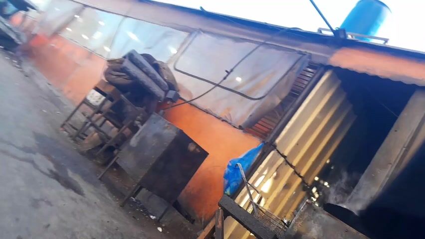 The video shows a street scene with a destroyed car, possibly after a fire or accident, alongside an orange building with partially visible windows. Two men walk past the camera; one is wearing a red hoodie, and the other is wearing a black jacket. There's a metal frame with cushions on top, positioned near the damaged car. The sky is visible in the background, suggesting it is daytime. Electrical wires run along the top of the building.