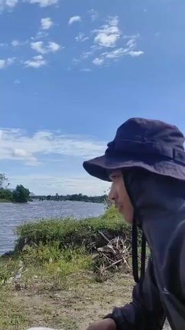 A person wearing a dark bucket hat and hooded top sits on the ground looking towards a wide river. The river has some debris, including a piece of wood, near the shore. The background features distant trees and hills under a partly cloudy blue sky.