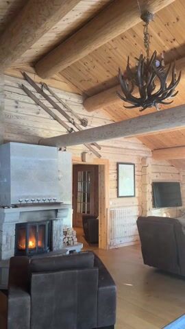 The video shows the interior of a rustic log cabin. The focal point is a stone fireplace with a roaring fire inside. Above the fireplace, on the mantel, are several small candle holders. Stacks of firewood are beside the fireplace. Two brown leather sofas sit in the room. A chandelier made of antlers hangs from the wooden ceiling. Old-fashioned skis are mounted high on the wall above the fireplace. The walls are constructed from wooden logs, and there's a wooden floor. A framed picture hangs on the wall, next to a baby gate blocking a doorway. A television is visible on the wall in the background. The room is well-lit, and the fire provides a warm glow. The camera slowly pans across the scene.