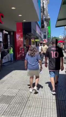 The video depicts a bustling city street scene on a sunny day. The camera follows a couple walking along a tiled sidewalk lined with various shops and vendors. The man is wearing a black Messi jersey with the number 10, paired with denim shorts and black sneakers. The woman is wearing a light blue shirt and a light-colored skirt, along with white sneakers. Other pedestrians can be seen strolling along the sidewalk in both directions. There are shops and buildings on both sides of the street, with a bus visible in the distance. A vendor is seen standing by a small cart displaying various trinkets. The atmosphere appears lively and urban.