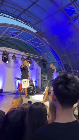 This is a short video clip showing a live musical performance. Two male performers are on a stage under a blue, curved roof structure. One performer, on the left, is wearing a black t-shirt and white shorts and is holding a microphone. The other performer, on the right, is wearing a dark shirt and is also holding a microphone and singing. In the background, there are stage lights and drum equipment. In the foreground, the backs of an audience are visible, with some individuals holding up their phones to record the performance. The atmosphere appears energetic, with one audience member raising an arm.