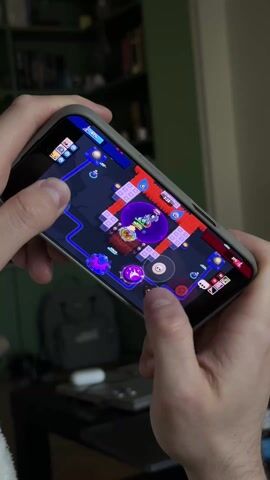 Here is a description of the video:

A person is playing a mobile game on their iPhone. The game appears to be a top-down shooter with colorful characters and a dark map with red pathways. The player is using both thumbs to control the character and aim/fire. The phone has a gray case. In the background, there's a dark green bookshelf, a gray backpack, and a laptop on a dark surface. The lighting in the room is somewhat dim. The shot is close-up, focusing on the phone and the person's hands.