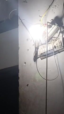 The video shows a white wall with a poorly mounted electrical panel, illuminated by a bright light bulb. There are multiple wires running from the panel, which appears to be old and worn. The wall has several holes and patches, suggesting it is in disrepair. To the left of the wall is a dark, undefined space, likely a doorway or corner of the room. The overall lighting is dim, with the exception of the area directly around the light bulb.