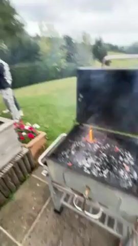 The video begins with a close-up of a grill with a smoky fire in the bottom. It is located on a patio with some flowers. A man is standing in the background. As the camera pulls back, it reveals a large grassy yard leading to a scenic view of rolling hills under a cloudy sky.