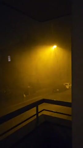 A porch railing is in the foreground, with a view of a street at night through a window or doorway. A single street lamp casts a yellow glow on the street, illuminating falling rain. There are a few cars parked on the street, and some buildings in the background. The overall scene is dark and appears to be during a heavy downpour.