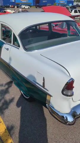The video pans from the rear passenger side of a white and teal classic car, possibly a 1955 Chevrolet Bel Air. The car is parked outdoors on an asphalt surface, with yellow lines visible on the ground. In the background, a large blue building with a yellow sign, resembling a retail store like Walmart, is present. Several other classic cars are also visible, including a red one with its hood open. People are milling around the parking lot. The camera moves forward, showing more of the car's side, including the door handle and a vent. The interior of the car, featuring a teal steering wheel and seats, is visible through the windows. The video continues to pan forward, focusing on the car's doors and windows, with the background elements remaining consistent. The scene appears to be a car show or a gathering of classic car enthusiasts.