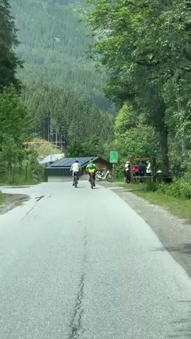 A group of people are outdoors on a paved road. Two people are on bicycles riding away from the camera. One is wearing a white shirt and the other is wearing a green vest. They are riding uphill towards buildings and a wooded mountain. There are several other bicycles parked near the buildings. A group of people are standing and sitting near the parked bicycles. The sky is overcast and the trees are green.