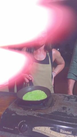 A woman in a black tank top and tan apron flips a green pancake in a black frying pan over a lit gas stovetop. She smiles after the flip, then places the pancake back in the pan. Two other people, both men, stand on either side of her, also wearing tan aprons. The stovetop is black and has a rusty appearance. The scene appears to be outdoors, at night.