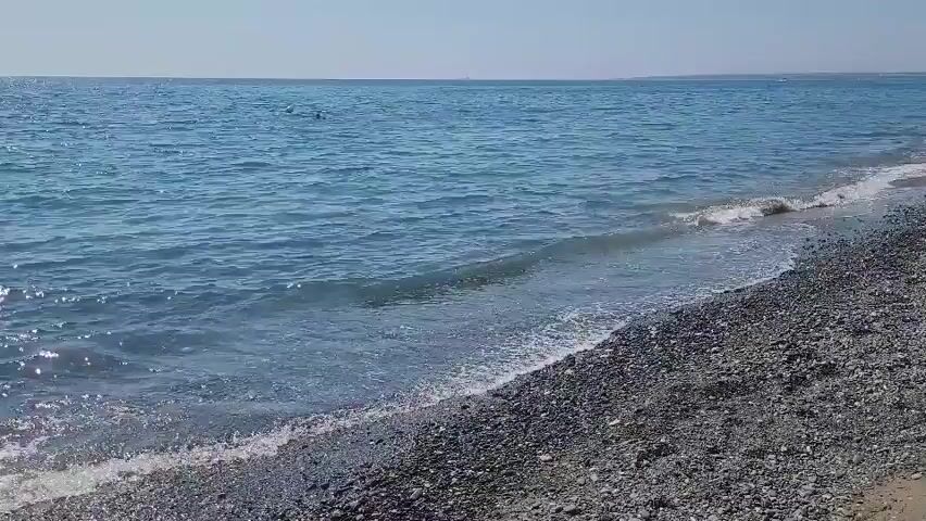 The video shows a body of water with small waves breaking on a shore composed of dark pebbles. The water is a clear blue and sparkles under what appears to be sunlight. The horizon is visible in the distance, with a faint line separating the sea from the sky, which is a pale blue. There are no people or identifiable objects in the frame, focusing solely on the natural landscape of the beach and the sea.