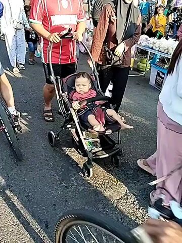 A man wearing a red and white striped shirt and black shorts, and a woman in a black hijab and brown top, are seen pushing a baby in a black stroller through a crowded outdoor market. The baby is wearing a red and black striped top and is seated in the stroller. A bicycle and a person's arm are visible in the immediate foreground. In the background, numerous people are walking and standing, alongside market stalls with umbrellas and goods. A large tree is also visible in the distance. The scene is illuminated by daylight.