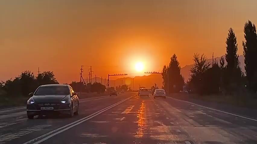 The video shows a view from a vehicle driving on a wet asphalt road at sunset. The sun is large and orange on the horizon, casting a warm glow over the scene. There are mountains in the distance, and trees line the sides of the road. Several cars are visible on the road ahead, also driving towards the sunset. There are power lines and utility poles visible on the left side of the road. The road has white lane markings. The overall impression is of a scenic drive during golden hour.