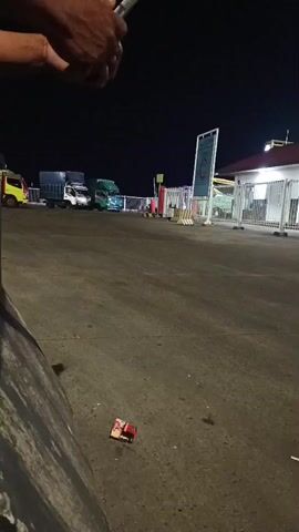 At night, an overhead shot shows a person's hands holding a phone, possibly recording. Below, a paved area with several trucks parked in the background. A lone cigarette pack sits on the ground in the foreground. The scene appears to be an outdoor area, possibly a rest stop or parking lot.