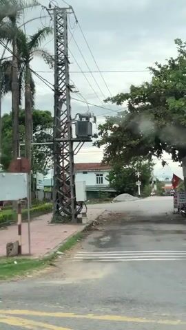 The video pans across a street scene in what appears to be Southeast Asia. On the left, a large billboard displays traffic safety information in Vietnamese, featuring various road signs and a cartoon depiction of a female traffic police officer. Beyond the billboard, palm trees and a building are visible. Further down the street, a Vietnamese flag waves. To the right of the billboard, a traffic sign indicating a U-turn is posted. The street has a sidewalk and grassy areas with hedges. Several buildings, including a house with a decorative facade, are visible along the street. People on motorcycles are seen riding past. A large truck is partially visible on the far right of the frame. The sky is overcast.