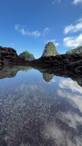 The video shows a still shot of a clear tide pool reflecting the bright blue sky with scattered white clouds. Beyond the pool, framed by dark, porous rocks, is a scenic ocean view featuring two small, green-topped islands and a partially visible coastline to the right. The water in the ocean appears calm and turquoise. The overall scene presents a tranquil and picturesque tropical landscape.