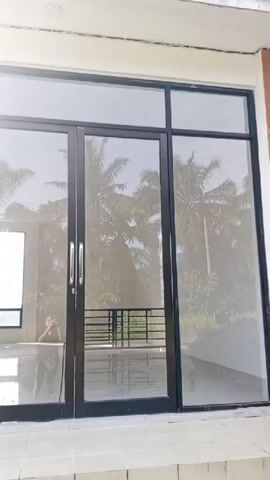 A reflection of a person is visible in a set of glass doors and windows, holding a phone and taking a selfie. The person is wearing a dark colored t-shirt and pants. In the background, there are palm trees and lush greenery. The scene appears to be outside a building with a light-colored tiled facade. The doors are dark framed and have silver handles. There is a small balcony visible through the glass with a metal railing. The lighting suggests it is daytime and sunny.