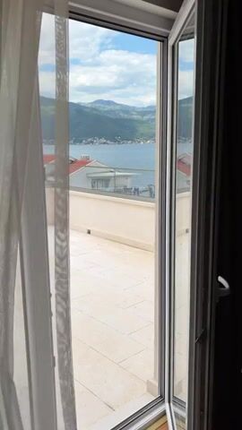 The video shows a tiled balcony with a white wall, through an open doorway. A sheer white curtain is visible to the left of the doorway. Beyond the balcony is a body of water, and in the distance there are mountains. There are houses on the shore. The sky is blue with clouds.